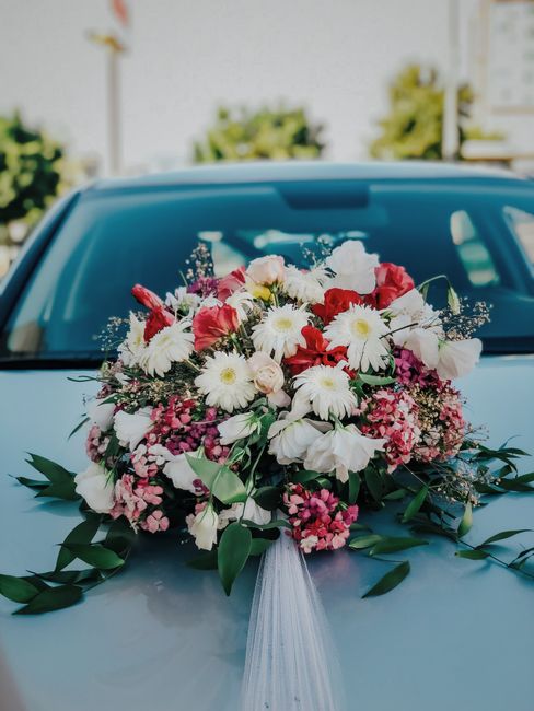 A vibrant floral arrangement featuring a mix of white daisies, pink and red roses, white lilies, and smaller pink and purple filler flowers, all lushly assembled into a rounded bouquet. The flowers ar