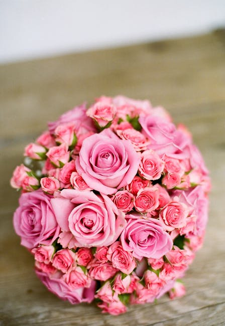 A tightly arranged bouquet of fresh pink roses and smaller pink spray roses, featuring soft, velvety petals and lush green stems, set on a rustic wooden surface. The flowers are bound together with mi