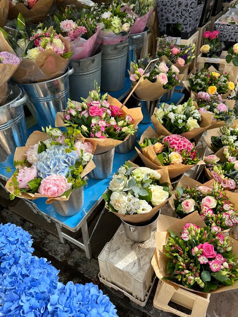 A vibrant display of freshly arranged floral bouquets and flower arrangements at a florist market stall, featuring a variety of pastel pinks, whites, blues, and greens. The flowers include roses, hydr
